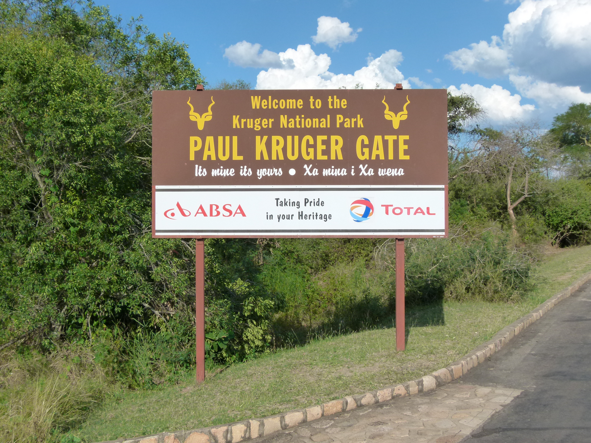 Bord voor de Paul Krugerpoort, Kruger National Park, bruin en wit met tekst en logo's, geplaatst in een graslandschap.
