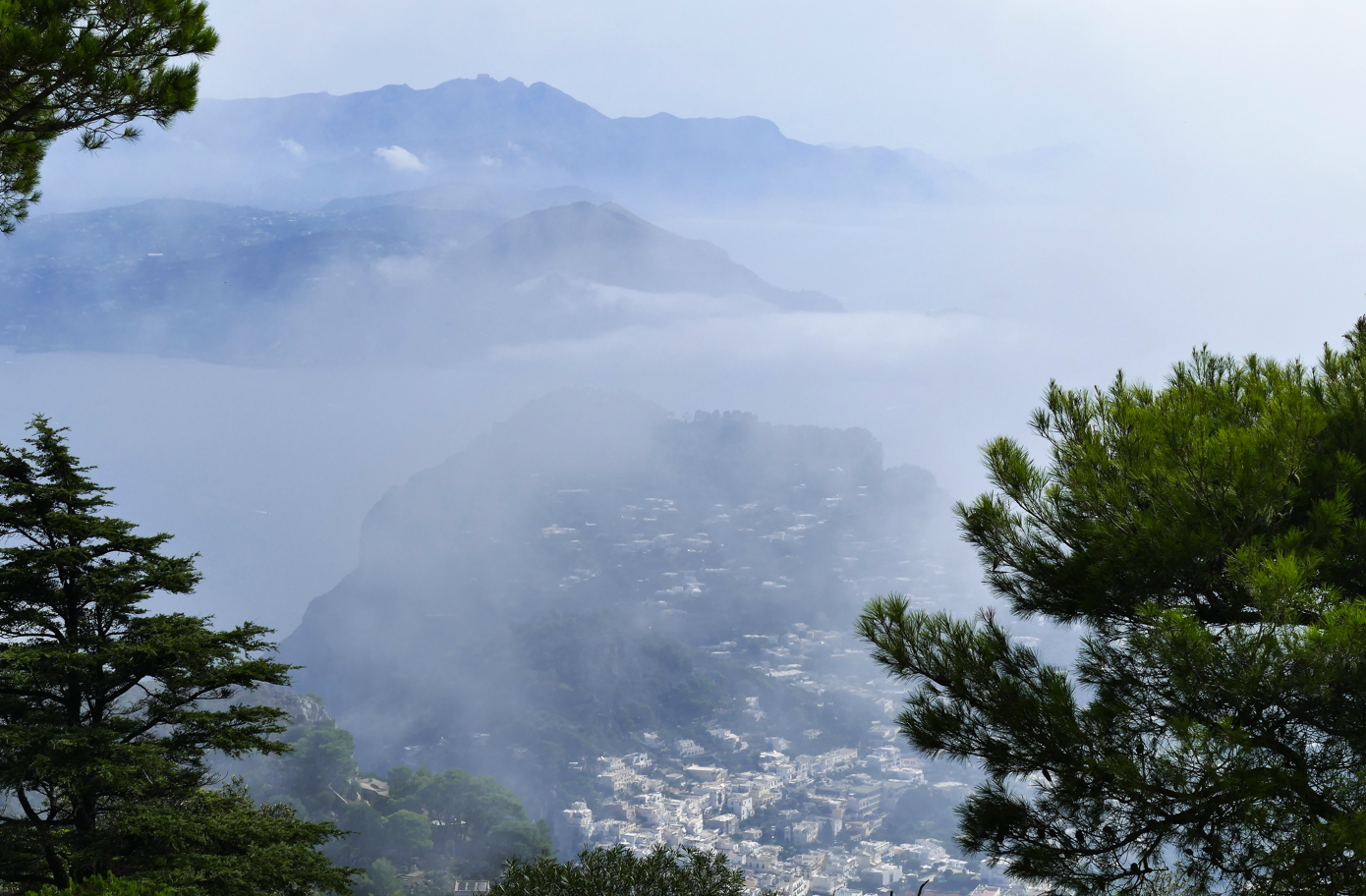 Uitzicht op Capri dat gedeeltelijk door mist aan het zicht onttrokken is, met bergen in de verte en bomen op de voorgrond.