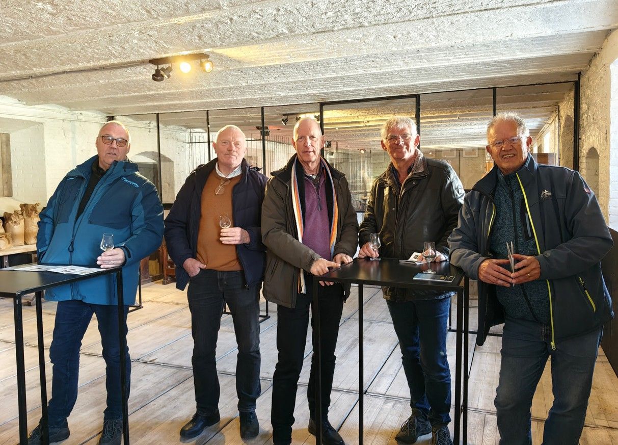 Vijf mannen staan in een rustiek, goed verlicht interieur en poseren voor een foto rond hoge bartafels.