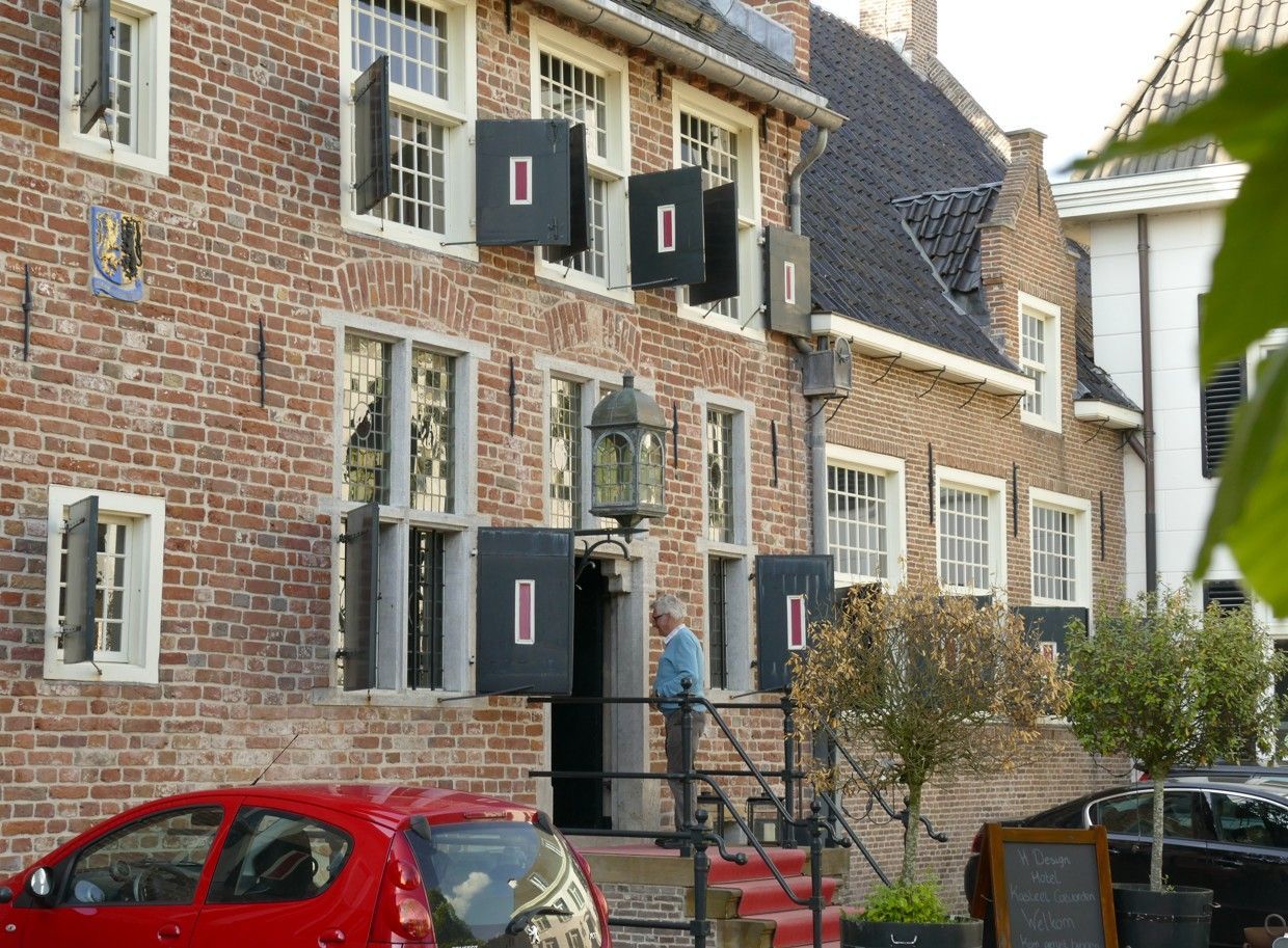 Rood bakstenen gebouw met zwarte luiken en een rode auto. Een man staat bij de ingang.