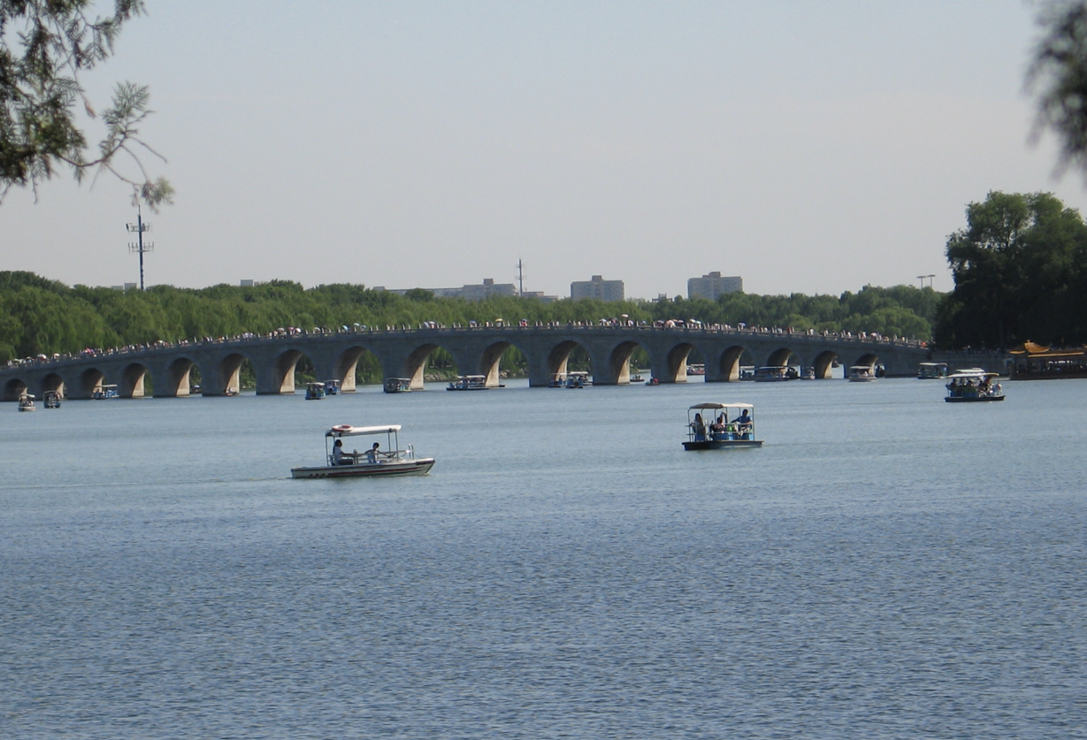 Een stenen boogbrug over een meer met boten. Mensen op de brug en in de boten; bomen en gebouwen op de achtergrond.