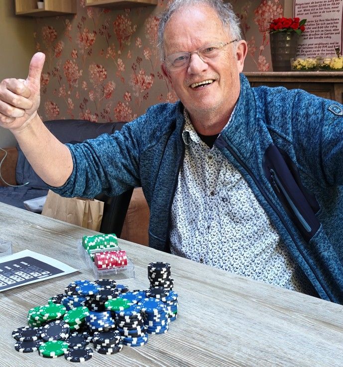 Een glimlachend persoon met een bril steekt zijn duim omhoog achter een tafel vol gekleurde pokerfiches.