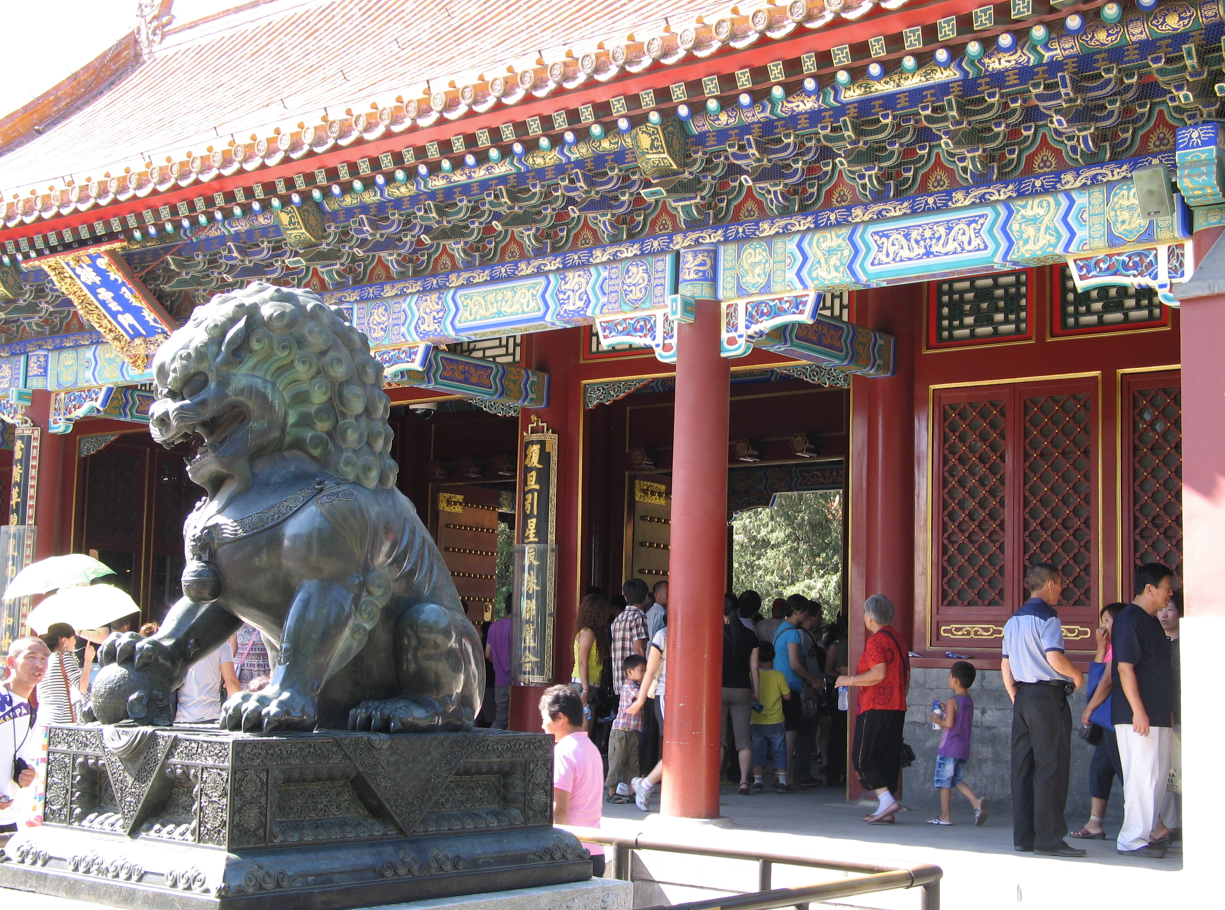 Een bronzen leeuwenbeeld bewaakt de ingang van een kleurrijk, rijk versierd Chinees gebouw; mensen staan ​​er vlakbij.