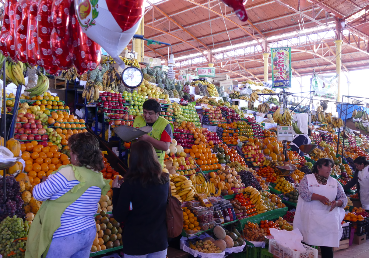 Fruitmarkt; diverse kleurrijke fruitstapels met winkelende mensen en een verkoper.