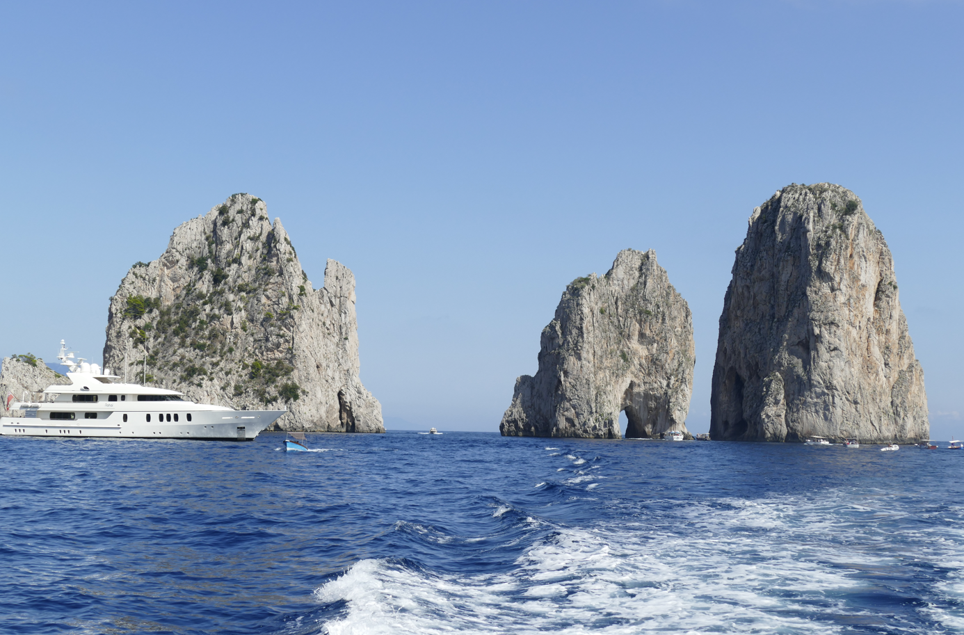 Een witte jacht vaart in de buurt van de Faraglioni-rotsen voor de kust van Capri, Italië; blauwe zee en lucht.