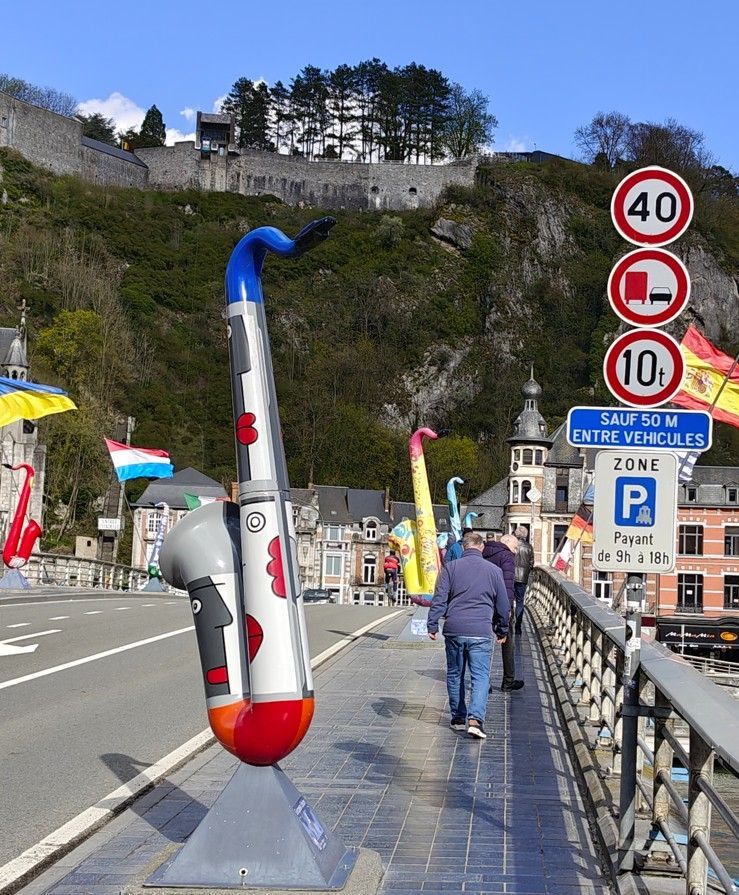 Kleurrijke saxofoonsculpturen sieren een voetgangersbrug in Dinant, België, onder een zonnige hemel met een rotsachtige klif.