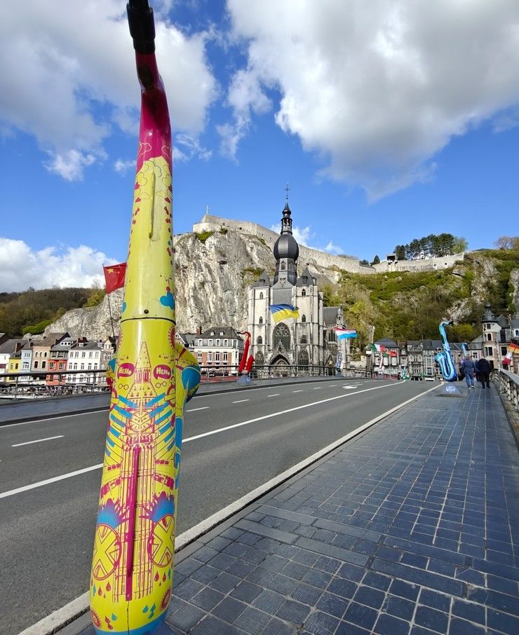 Een kleurrijk, kunstzinnig versierd saxofoonbeeld staat op een brug voor een historische kerk en een klif in Dinant.