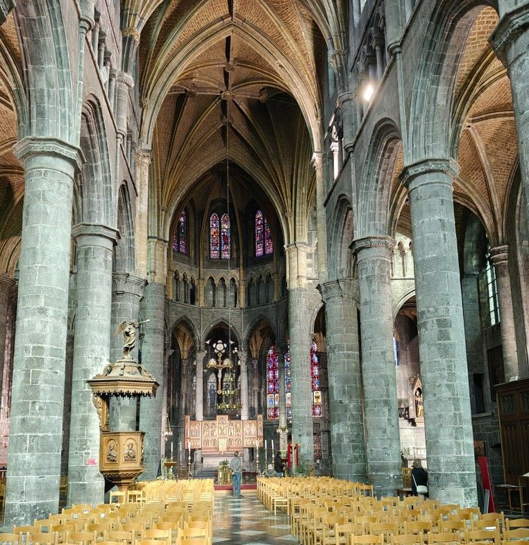 Interieur van een grote gotische kerk met stenen pilaren, gewelfde plafonds, glas-in-loodramen en rijen houten stoelen.