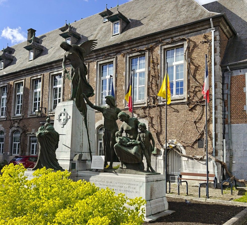 Een bronzen oorlogsmonument met figuren voor een bakstenen gebouw met vlaggen op een stadsplein onder een zonnige hemel.