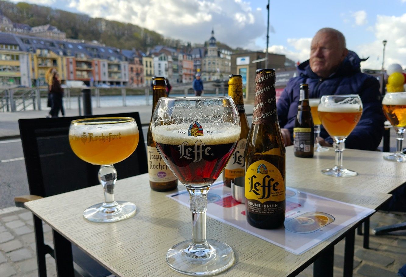 Een tafel met flessen en glazen Leffe-bier op een terras aan de rivier, met een persoon op de achtergrond.