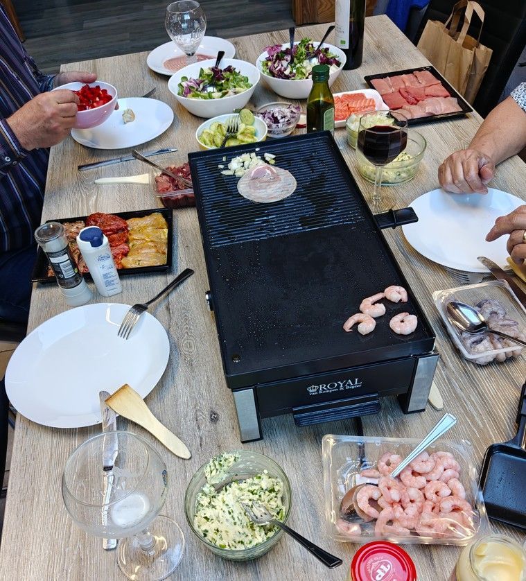 Een tafelgrill wordt omringd door borden, kleine schaaltjes met salades, garnalen en kruiden voor een gezamenlijke maaltijd.