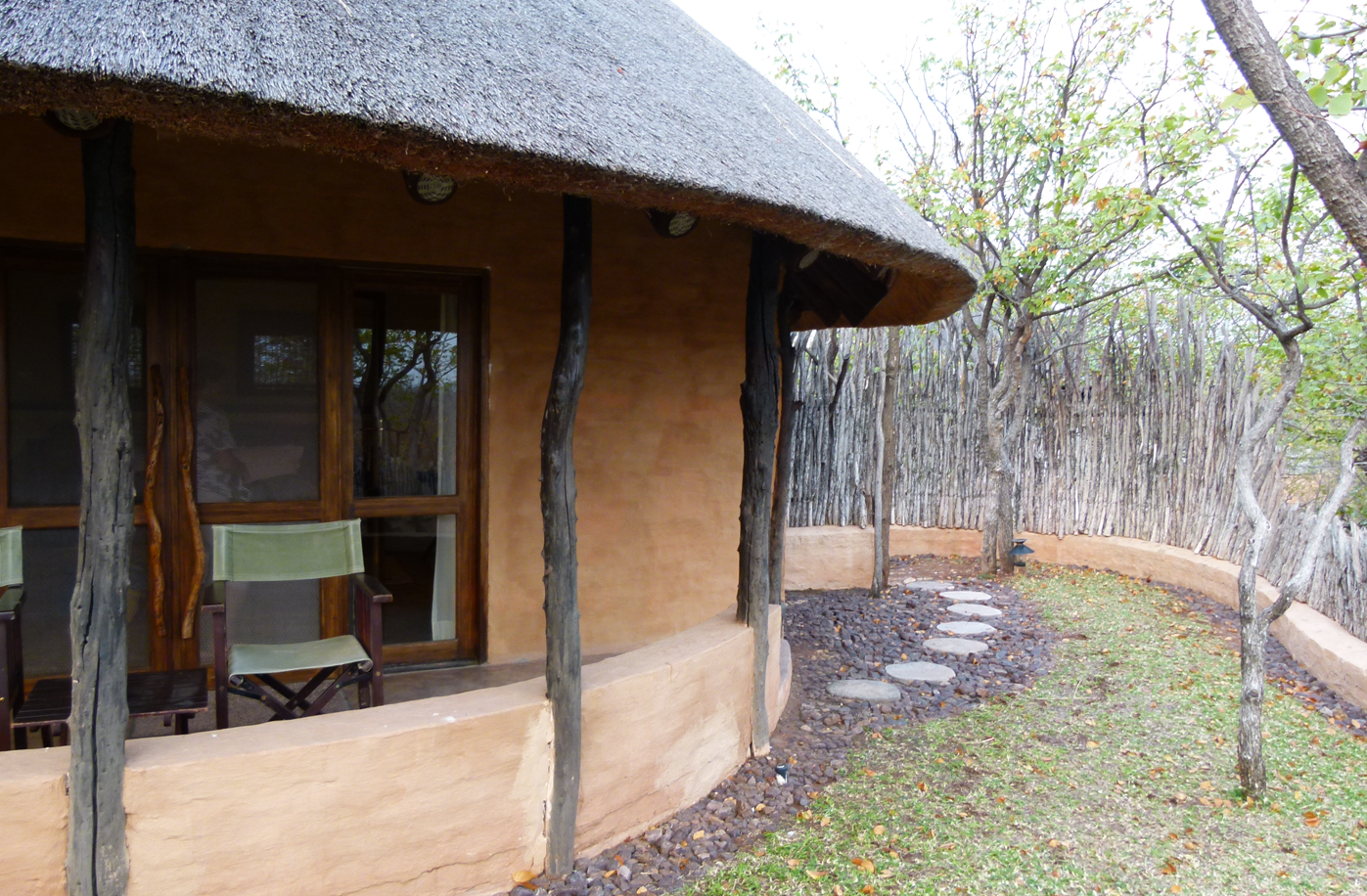 Een lodge met een rieten dak, bruine muren, houten pilaren en een stenen pad in een bosrijke omgeving.