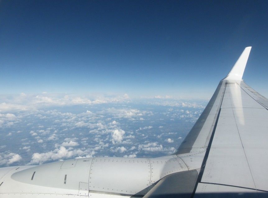 Uitzicht vanuit het vliegtuigraam, vleugel in beeld, over verspreide wolken, blauwe lucht.