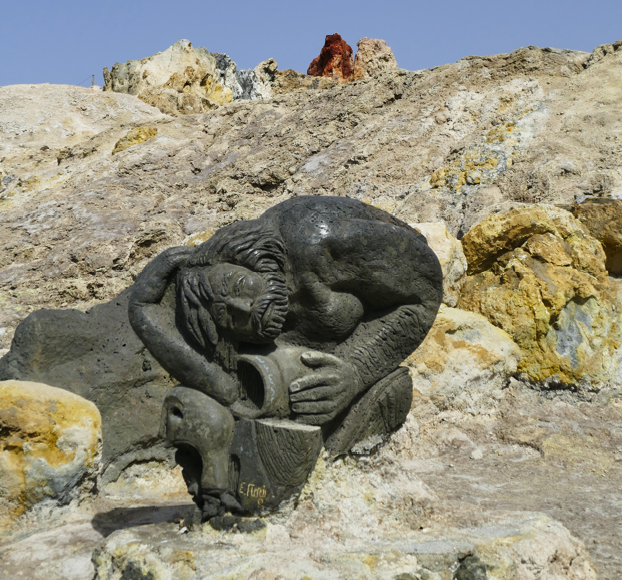 Een sculptuur van een persoon die een pot vasthoudt, tegen een achtergrond van zwavelhoudende, geelachtige rotsen.