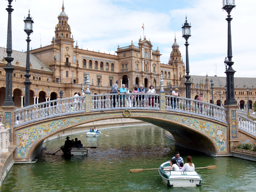 Mensen op een brug over een kanaal met roeiboten voor een sierlijk gebouw in Sevilla, Spanje.