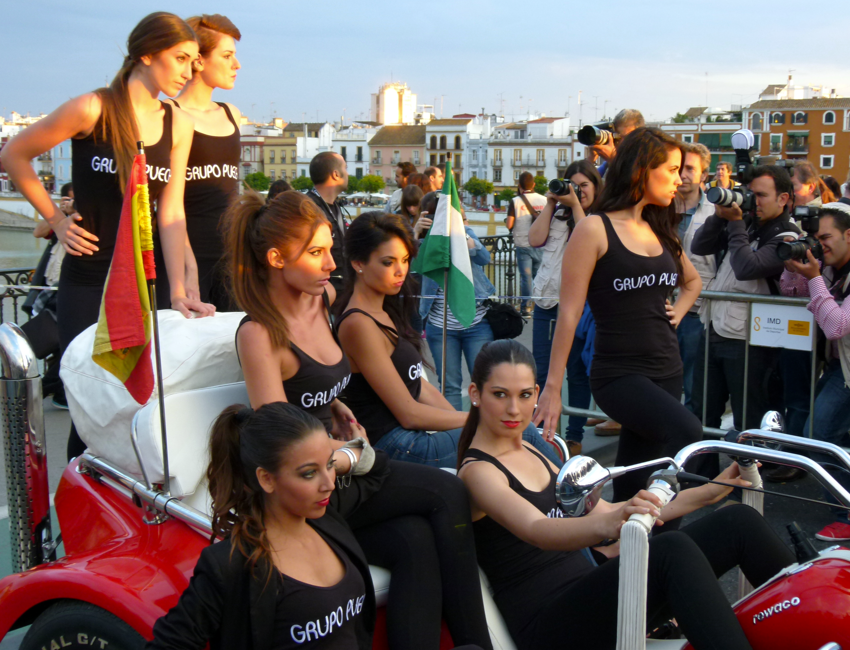 Vrouwen in een rode auto, gekleed in zwarte topjes met logo, poseren voor foto's. Op de achtergrond Sevilla