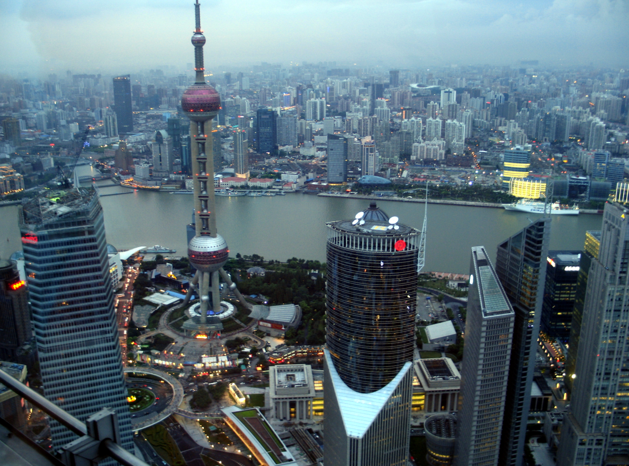 De skyline van Shanghai bij zonsondergang, met de Pearl Tower en de rivier; veel hoge gebouwen verlicht.