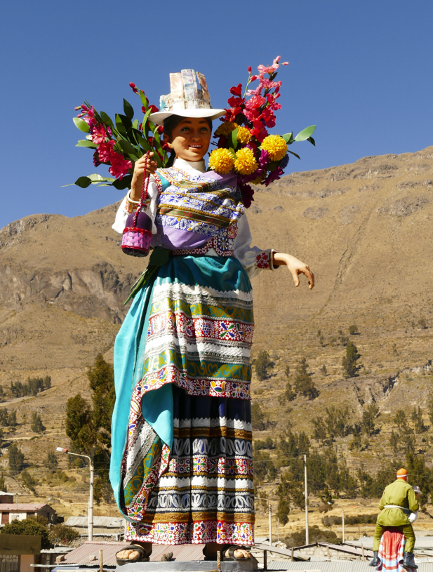 Standbeeld van een vrouw in traditionele kleding, met bloemen in haar hand. Berglandschap op de achtergrond, blauwe lucht.