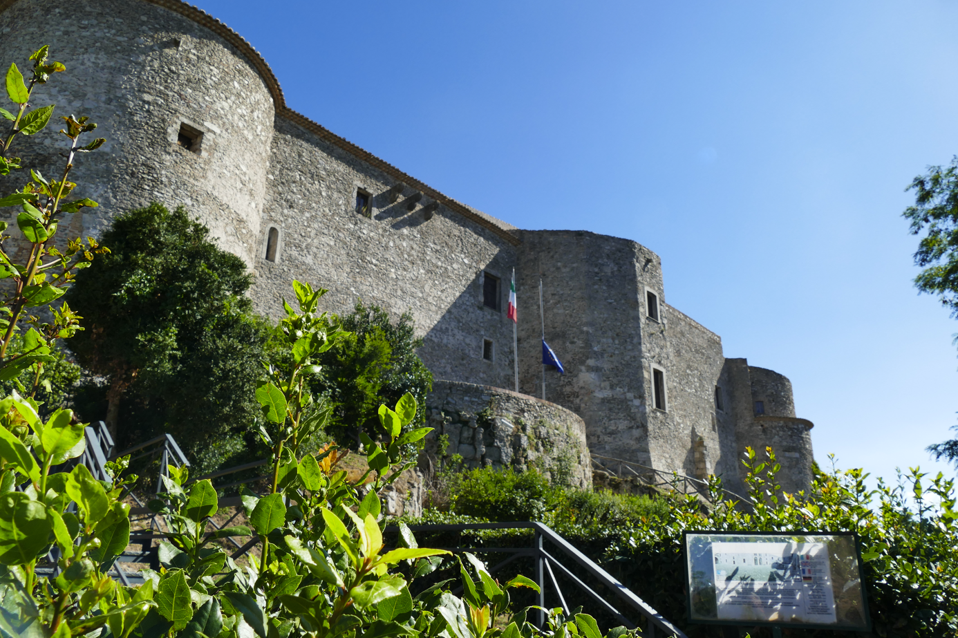 Stenen kasteel met ronde torens en de Italiaanse vlag onder een helderblauwe hemel.