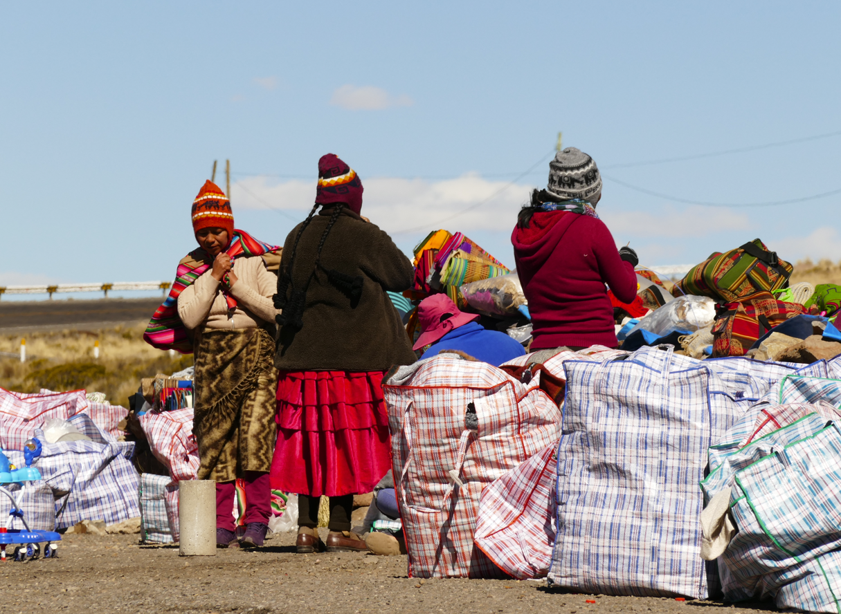 Vrouwen in kleurrijke kleding verkopen goederen vanuit grote tassen langs de weg.