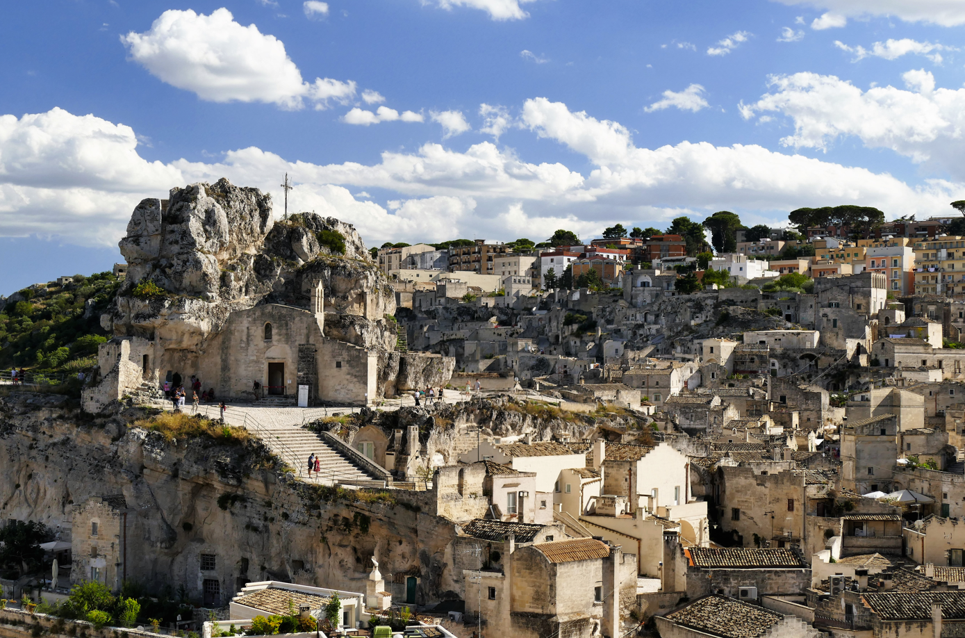 De Italiaanse stad Matera, gebouwd tegen een rotsachtige heuvel, met stenen gebouwen onder een blauwe hemel met wolken.