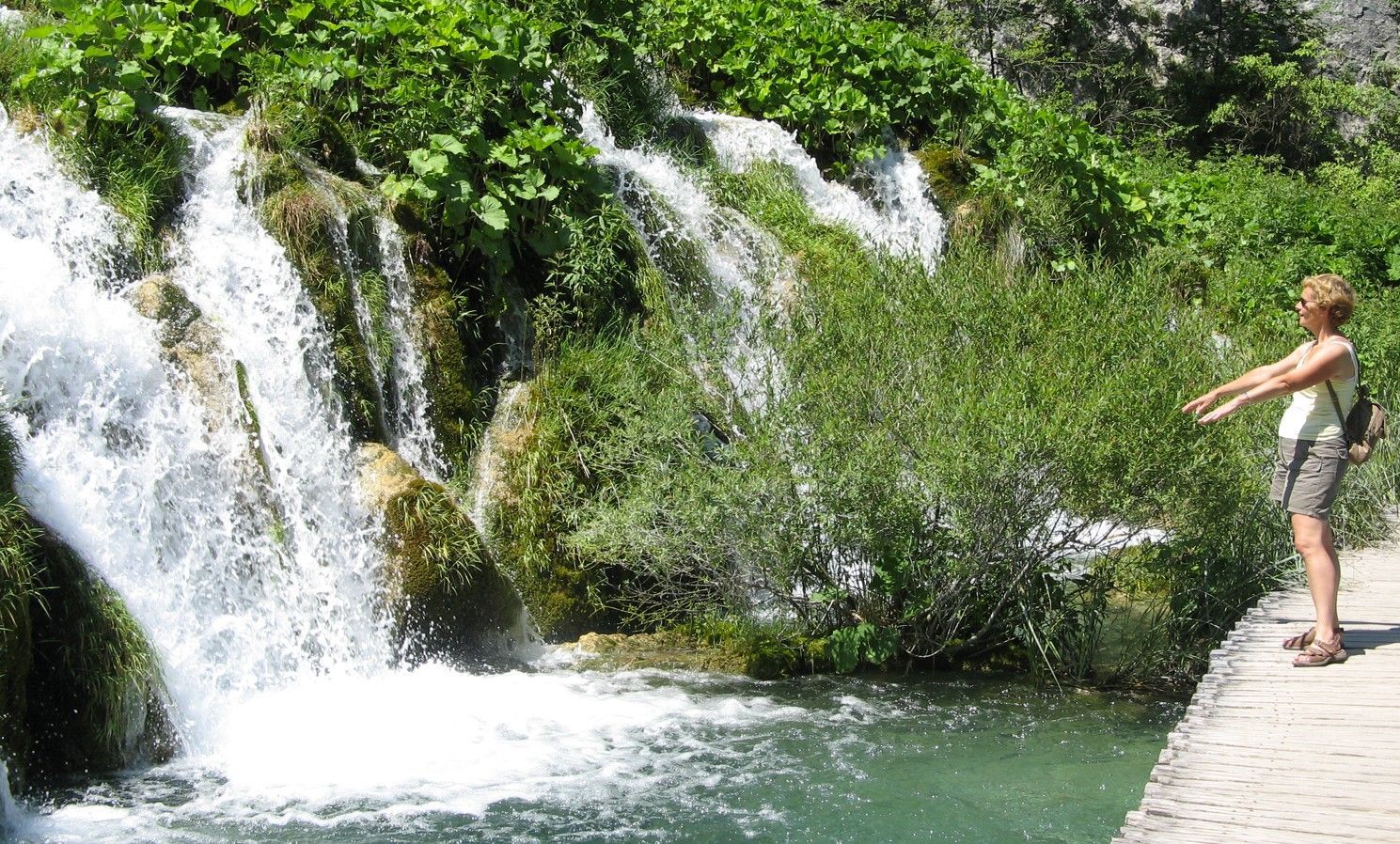 Een vrouw staat bij een waterval met uitgestrekte armen. Smaragdgroen water en weelderige begroeiing omringen een stenen pad.