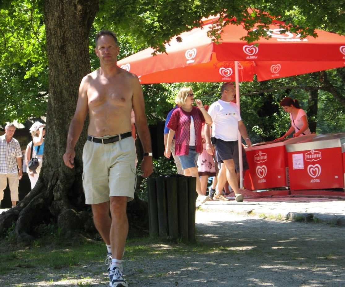 Een man in korte broek loopt naar de camera toe; op de achtergrond staan ​​mensen en een ijsje onder een rode paraplu.