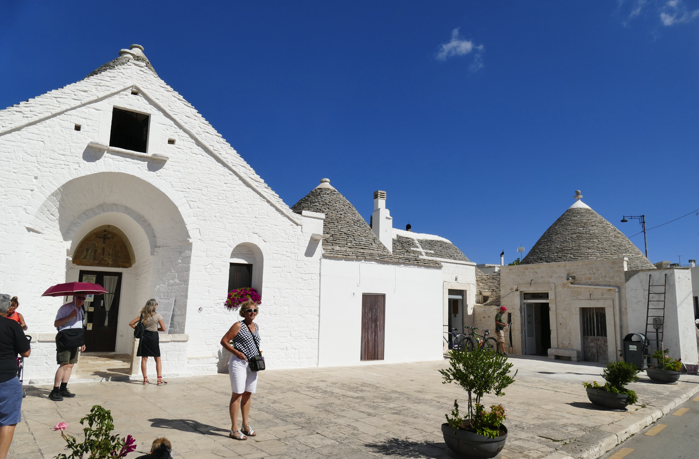 Witte trulli-gebouwen met kegelvormige daken in Alberobello, Italië, onder een blauwe hemel, met toeristen.