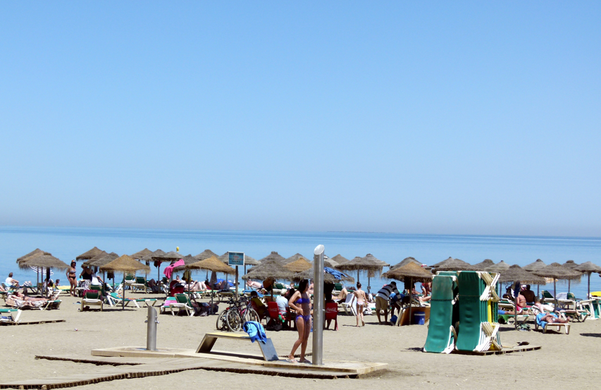 Strandtafereel met rieten parasols, zonnebadende mensen en de oceaan onder een blauwe hemel.