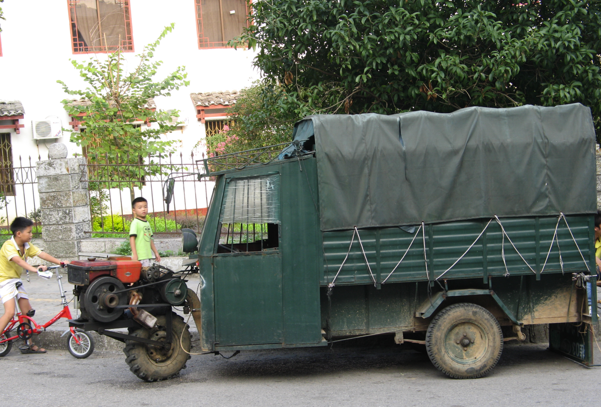 Groene gemotoriseerde driewieler met laadruimte; twee jongens op de stoep, waarvan er één een kleine rode fiets duwt.