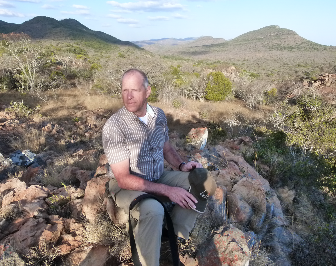 Een man zit op rotsen in een droog, heuvelachtig landschap; hij draagt ​​een overhemd met een patroon en een kaki broek