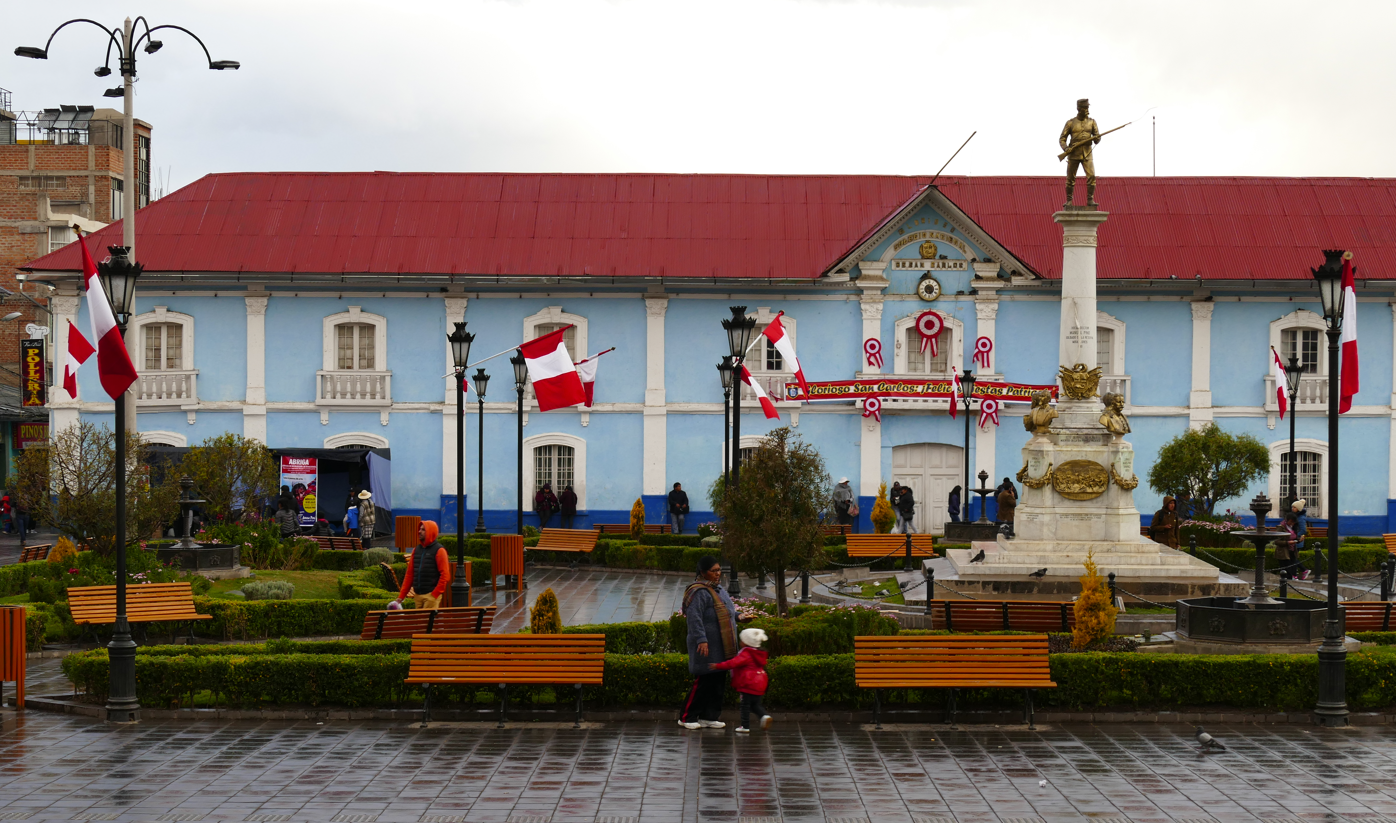 Een blauw gebouw met een rood dak, Peruaanse vlaggen en een monument op een plein. Mensen wandelen er rond.