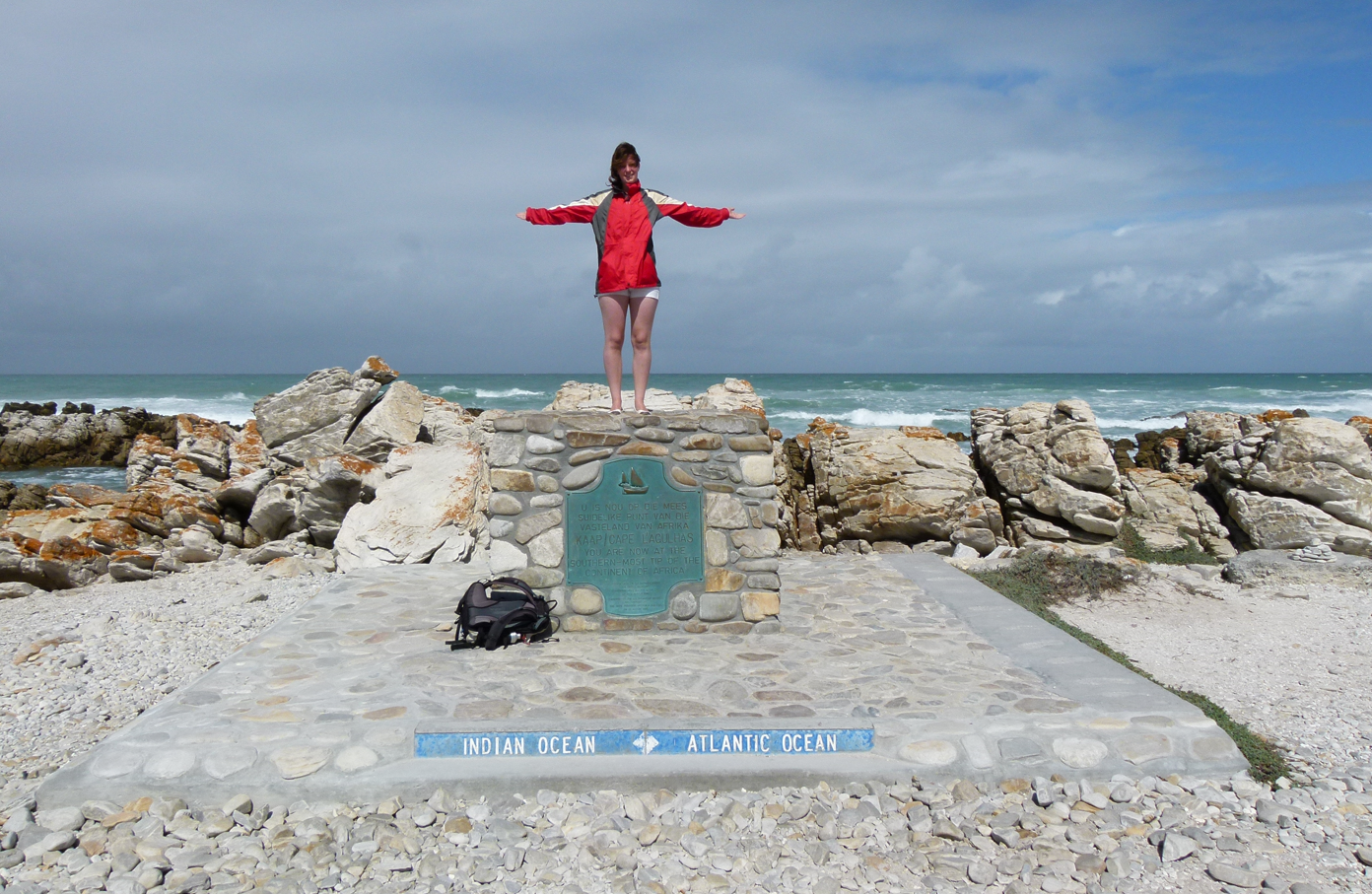 Vrouw staat op een stenen monument, met uitgestrekte armen, vlakbij de oceaan, onder een bewolkte hemel.