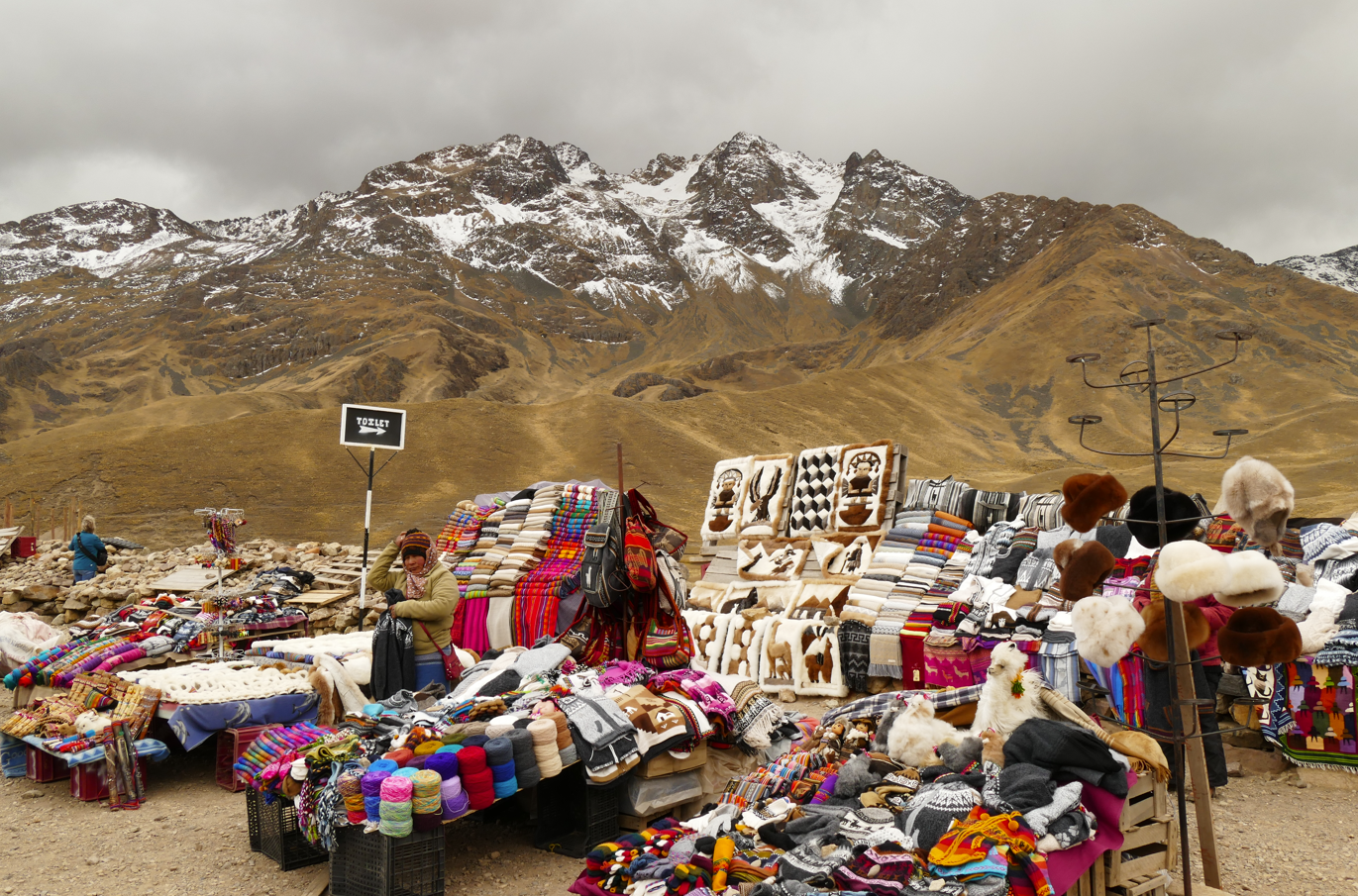 Een openluchtmarkt met kleurrijke textielwaren, sieraden en hoeden tegen een achtergrond van besneeuwde bergen. 