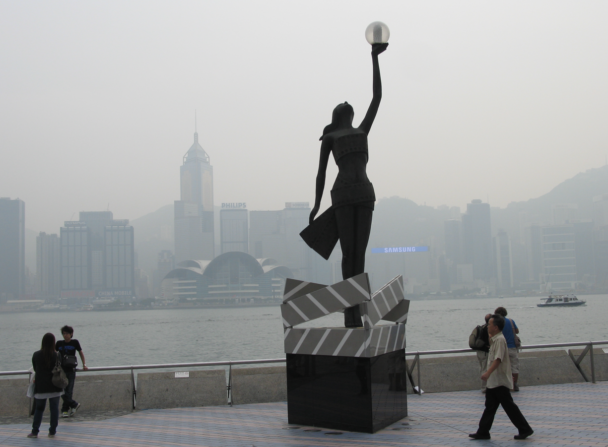 Standbeeld van een vrouw met een bol aan de waterkant; de skyline van Hongkong op de achtergrond.