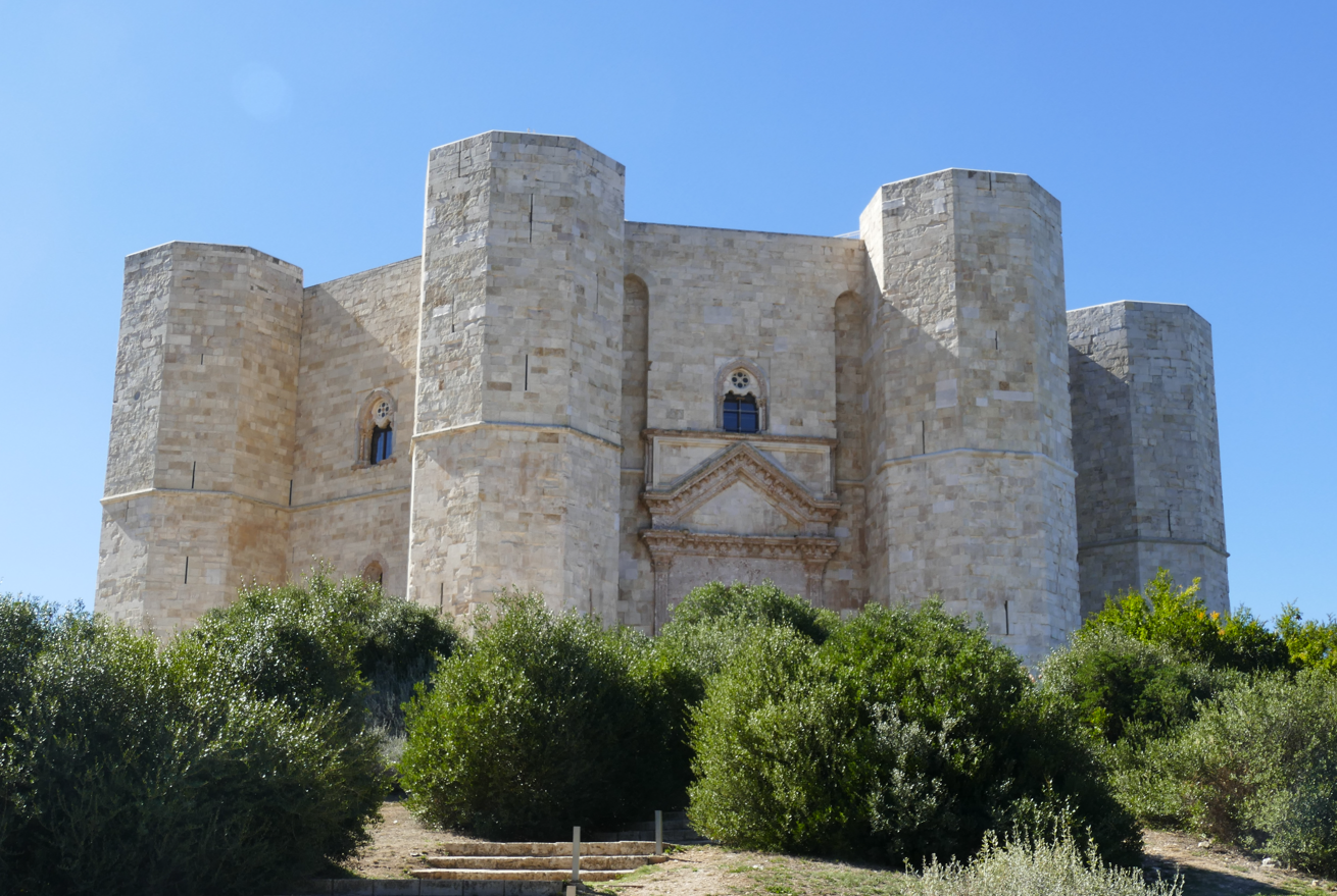 Castel del Monte, Italië, vesting met achthoekige torens. Lichte stenen tegen een blauwe lucht, struikgewas op de voorgrond.
