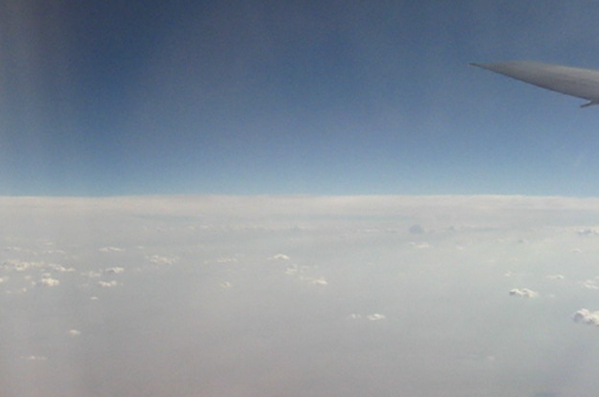 Uitzicht vanuit een vliegtuigraam, met een laag wolken onder een helderblauwe hemel, en een zichtbare vleugeltip.