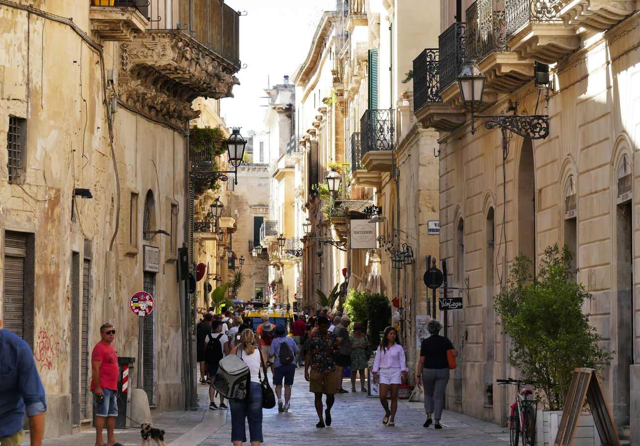 Een smalle straat in een Italiaans stadje, mensen lopen erlangs. Beige gebouwen met balkons en planten sieren de straat.