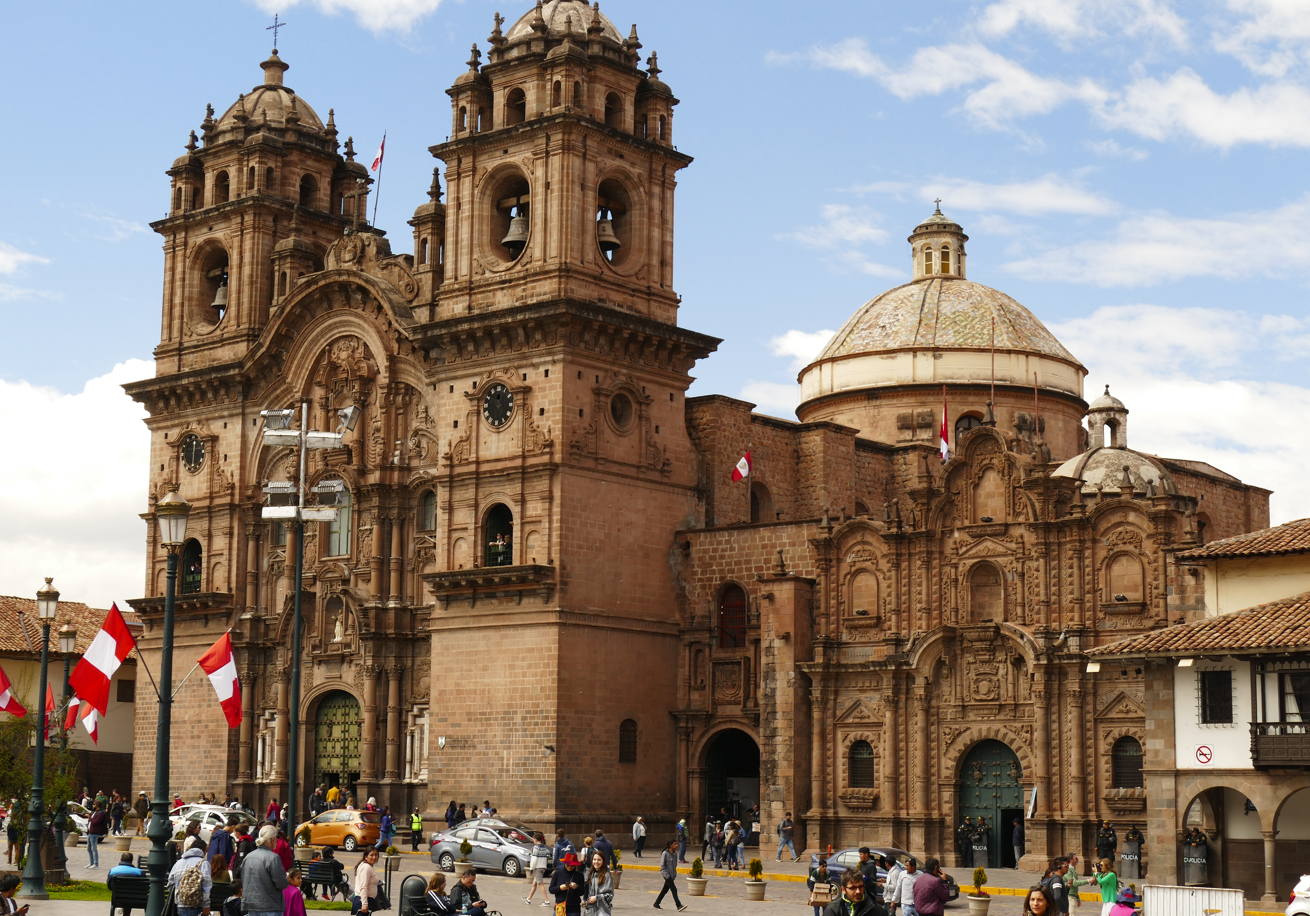 De buitenkant van de kathedraal van Cusco in Peru, een grote bruine stenen kerk met klokkentorens, een koepel en vlaggen.