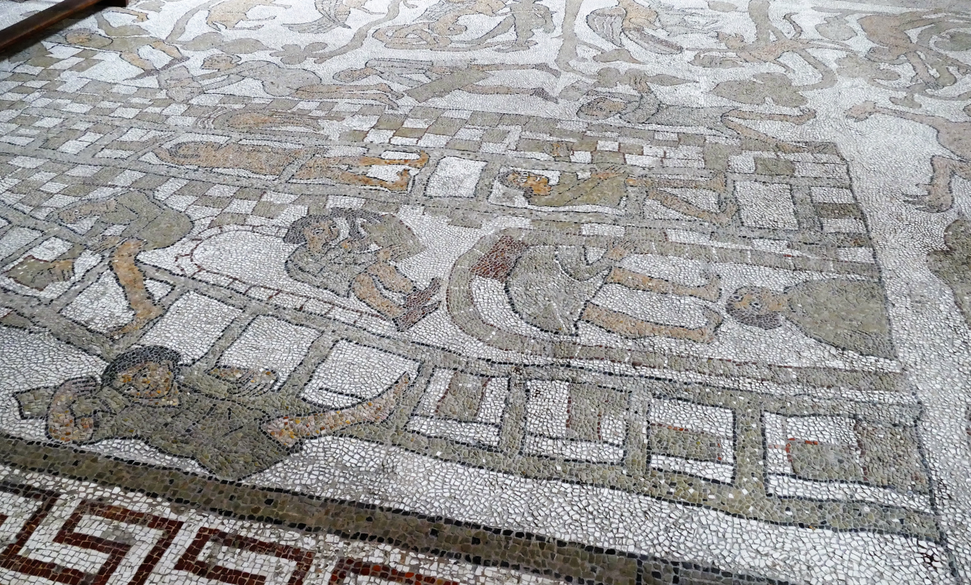 Oude mozaïekvloer met een gedetailleerd stadsgezicht; figuren, gebouwen en een decoratieve rand.