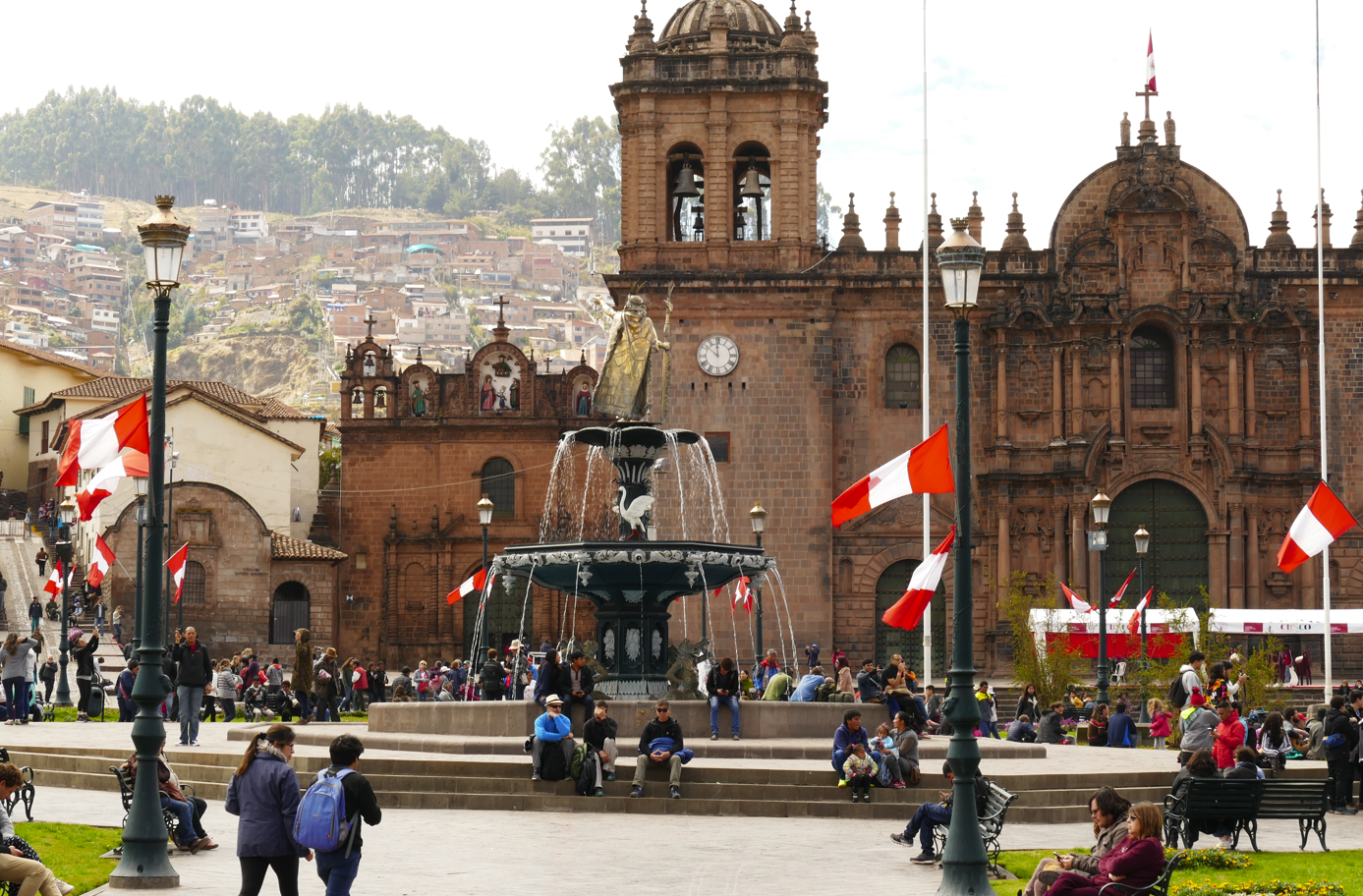 Plein in Cusco met sierlijke fontein en kerk, Peruaanse vlaggen wapperend, mensenmassa verzameld.