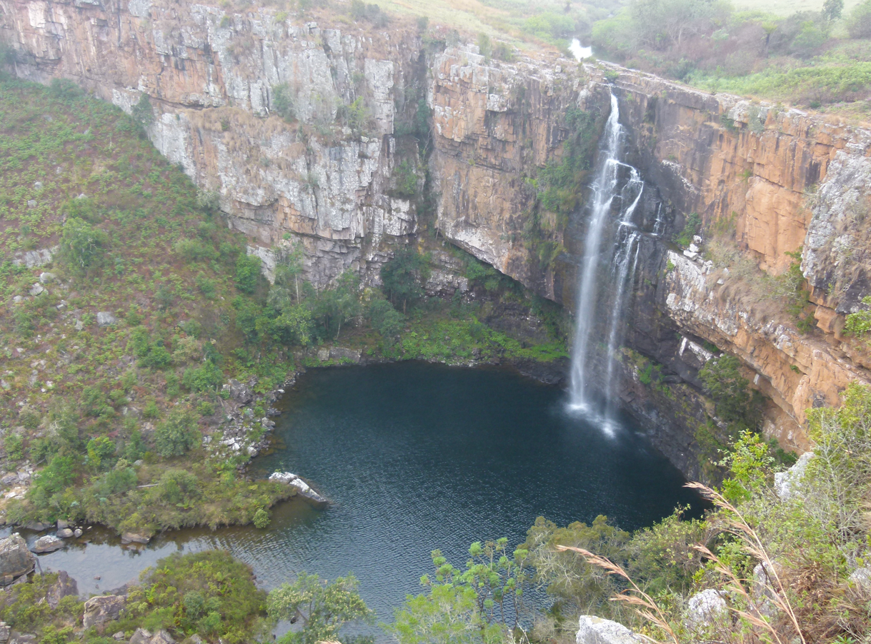 Een waterval die neerstort in een donkere, ronde poel, omgeven door rotswanden en schaarse begroeiing.