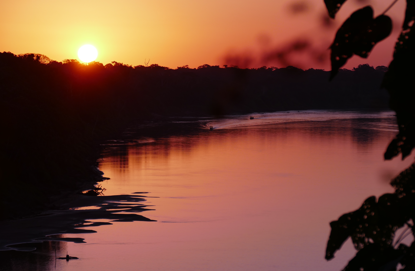 Zonsondergang boven een kalme rivier die de roze en oranje lucht weerspiegelt; silhouetten van bomen omlijsten het water.