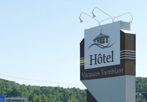 Het bord 'Hotel Vacances Tremblant' met logo en naam, tegen een blauwe lucht, vlakbij bomen.