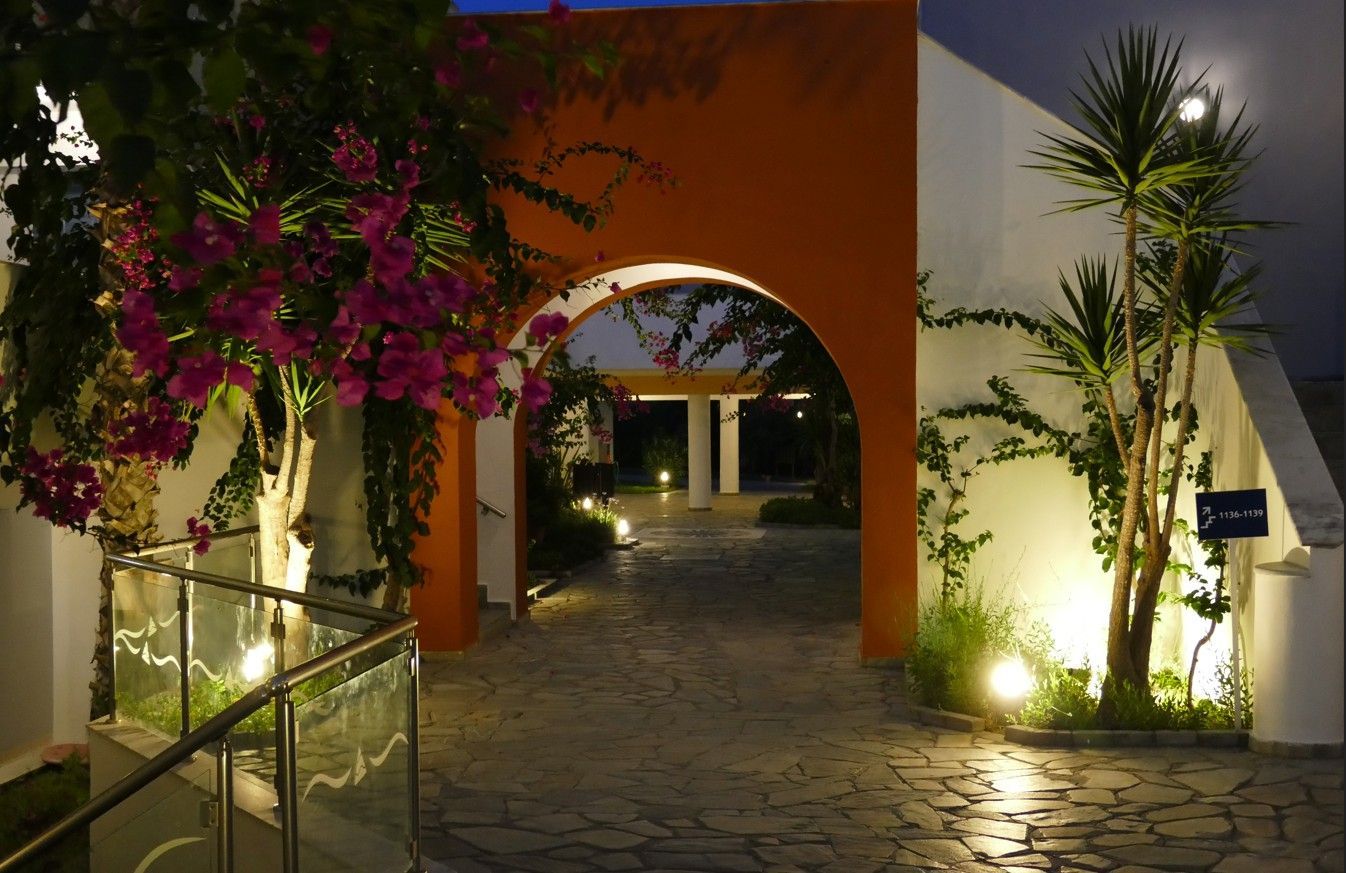Een oranje boog leidt naar een tuinpad, verlicht door grondlampen, met bougainvillea en palmbomen.