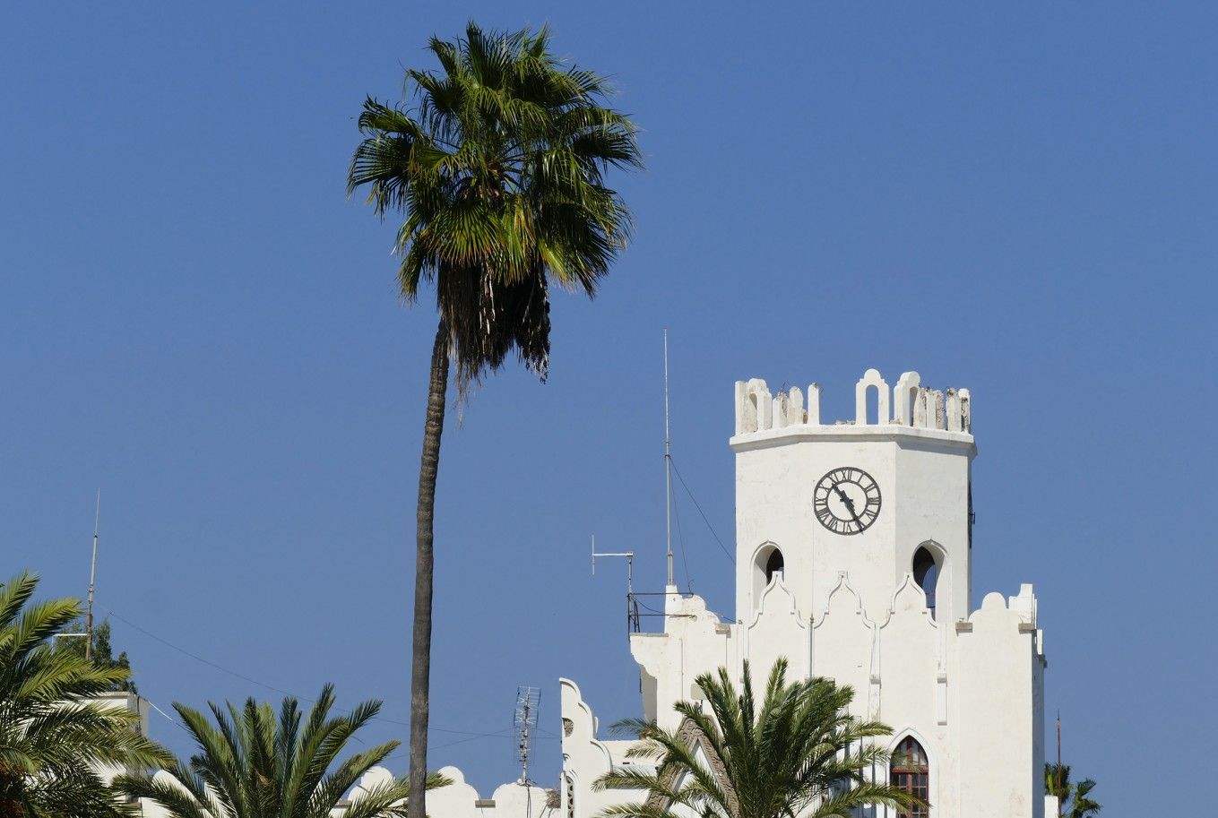 Witte klokkentoren met boogvormige kantelen en palmboom tegen een blauwe lucht.