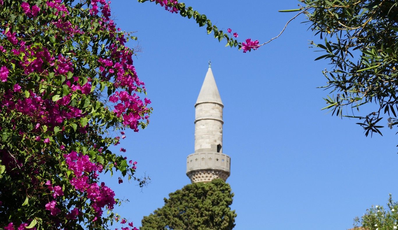 Stenen minaret met kegelvormige top, omlijst door roze bougainvillea en een blauwe hemel.