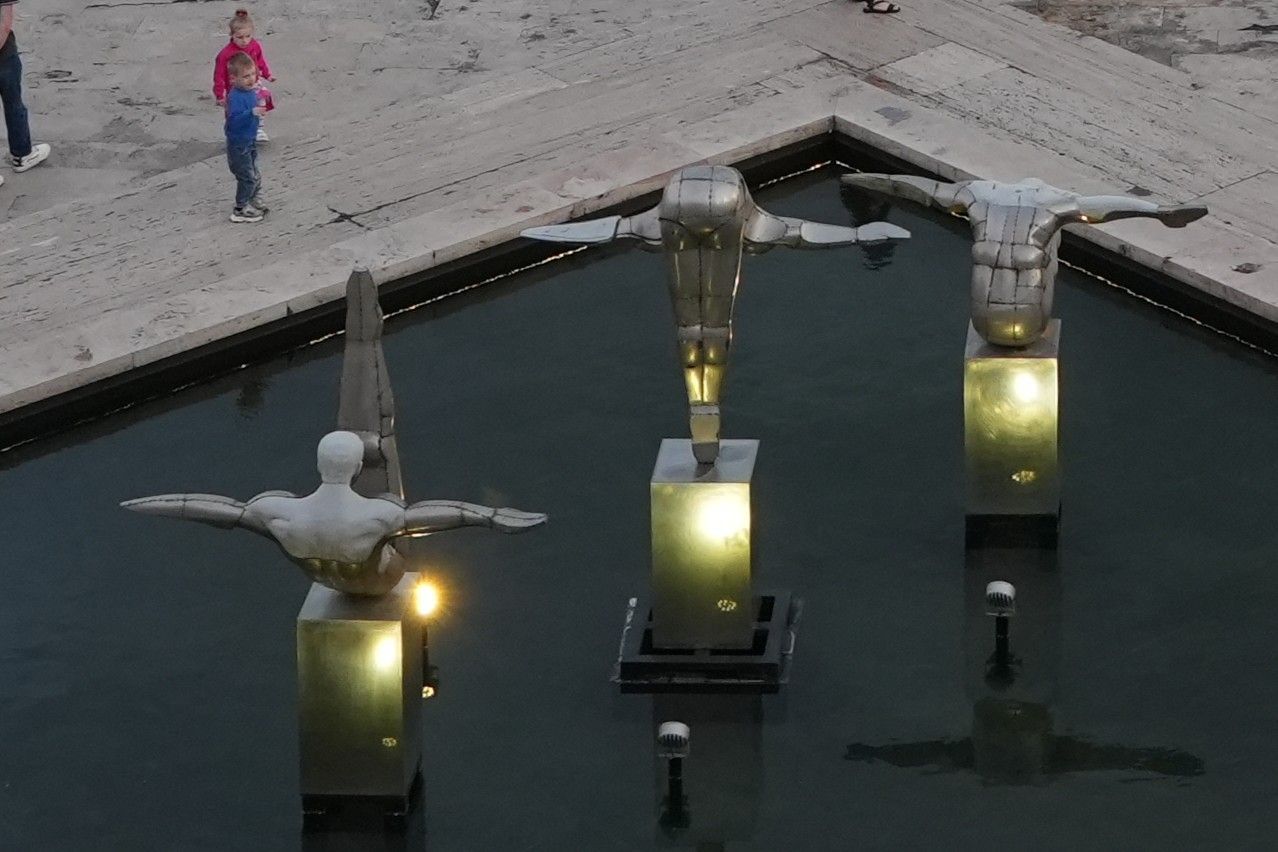 Vogelbeelden op verlichte sokkels in een donkere fontein, met een persoon die vlakbij over het stenen pad loopt.