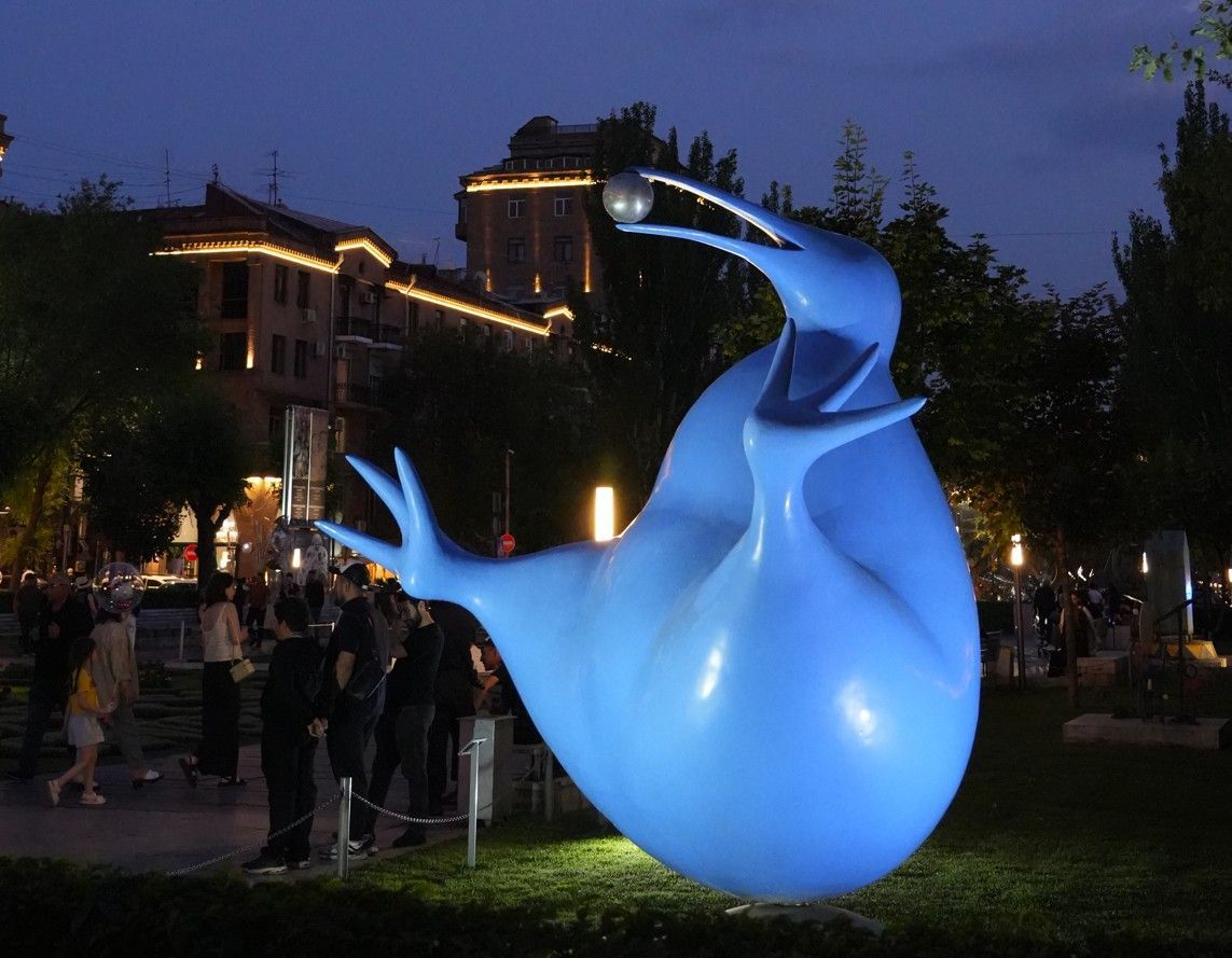 Een abstract, lichtgevend blauw sculptuur in een park 's nachts, met mensen eromheen en verlichte gebouwen op de achtergrond.