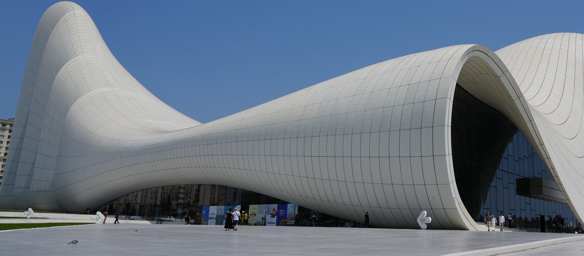 Een modern, wit gebouw met een gebogen vorm, een grote open ingang en mensen die buiten rondlopen.